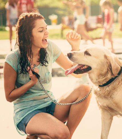 A lady with wet hair stoops to pet her dog, who has been playing at the Splash Pad.