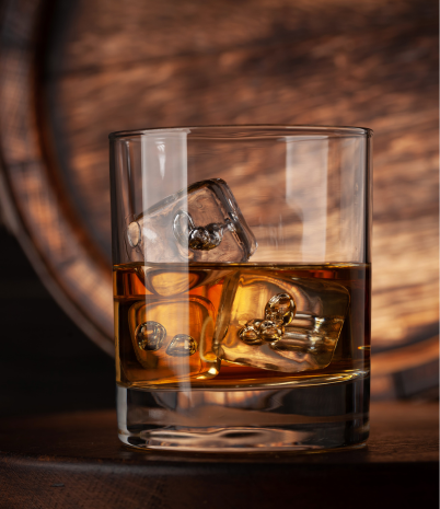 A glass of bourbon with ice sits in front of a wooden distillery barrel.
