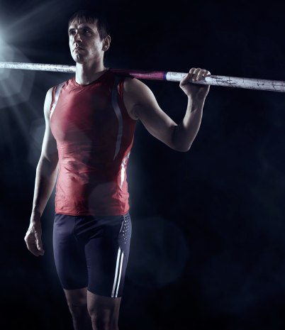 A pole vaulter stands against a dark background with a pole across the back of his shoulders.